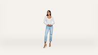 A woman stands against a plain white background, wearing a white long-sleeve top, light blue ripped jeans, and white heels.