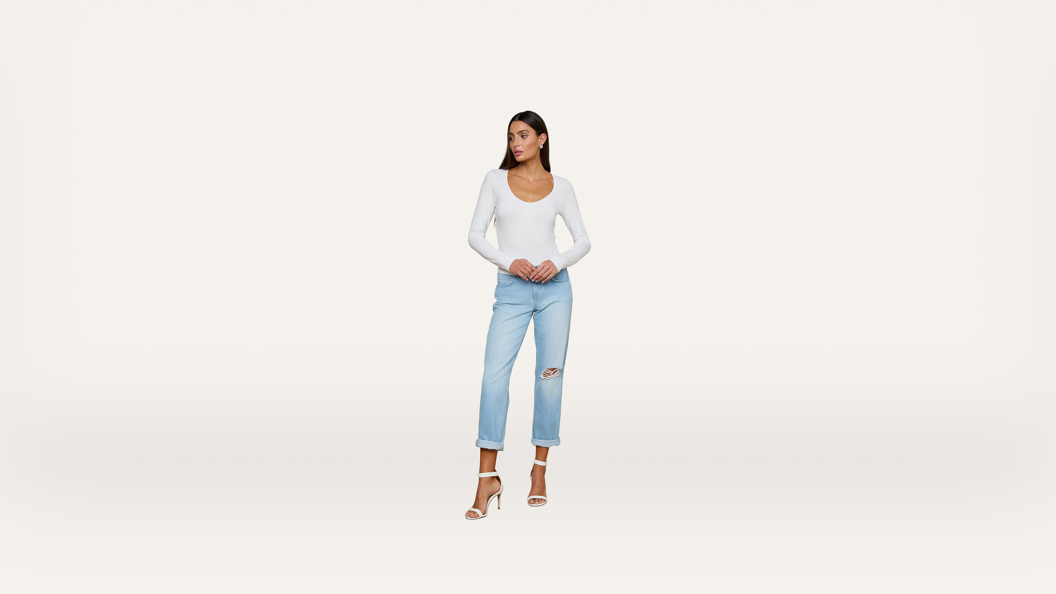 A woman stands against a plain white background, wearing a white long-sleeve top, light blue ripped jeans, and white heels.