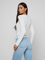 A woman with long brown hair, wearing the Cassandra Ruched Bodysuit—a white long sleeve top with a scoop neckline—paired with light blue jeans, stands sideways and looks over her shoulder against a plain, light background.