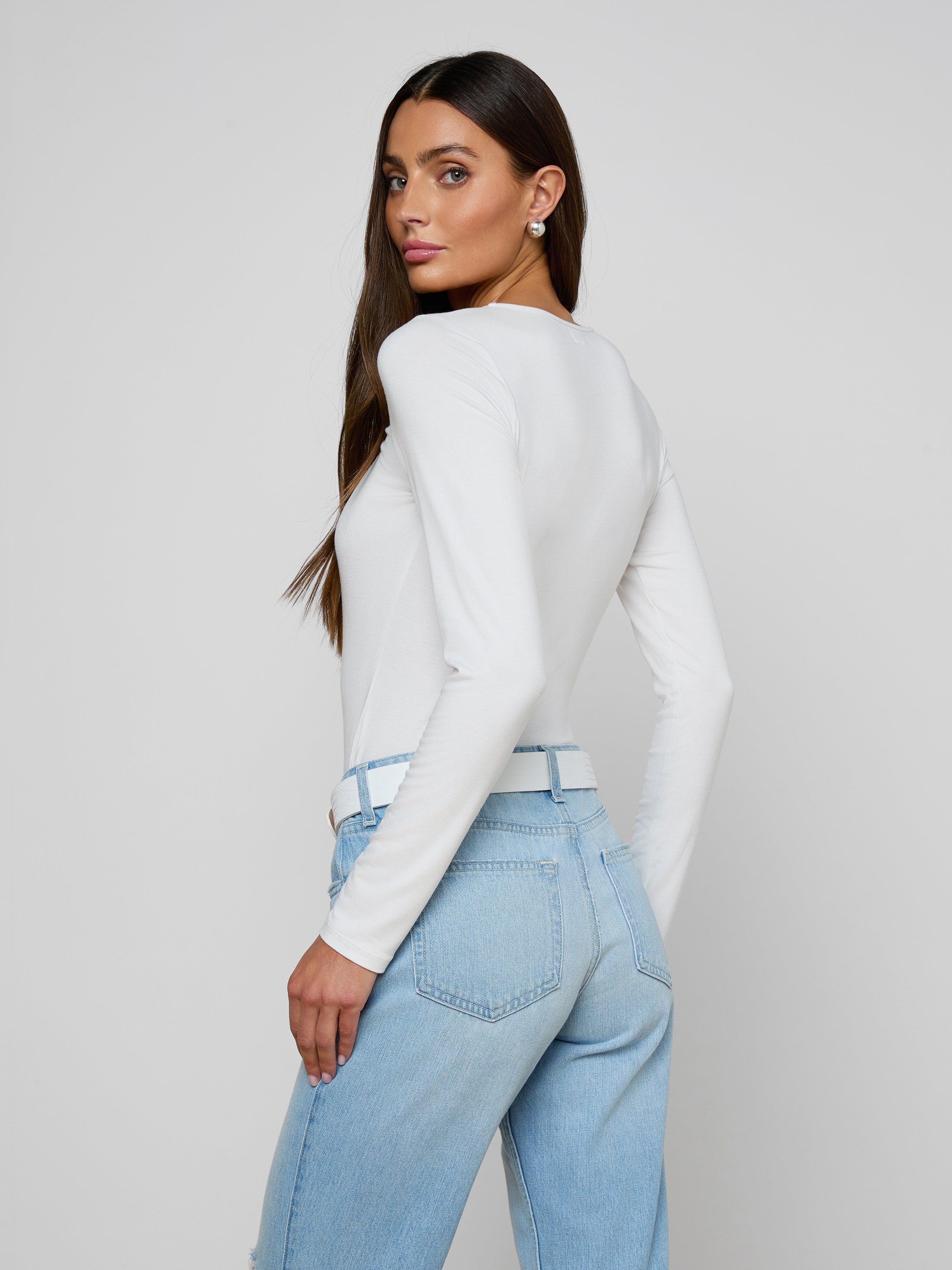 A woman with long brown hair, wearing the Cassandra Ruched Bodysuit—a white long sleeve top with a scoop neckline—paired with light blue jeans, stands sideways and looks over her shoulder against a plain, light background.