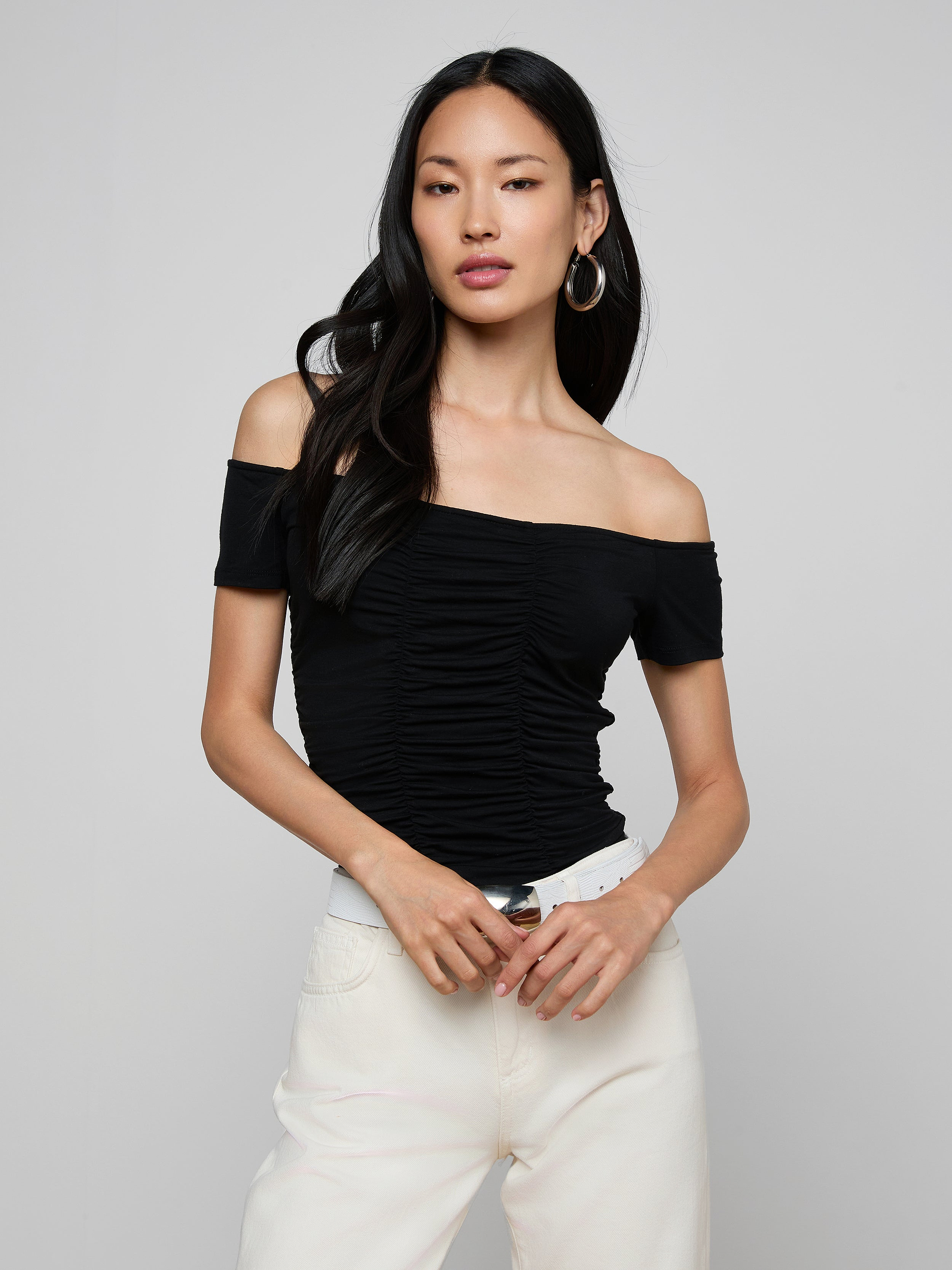 A woman with long dark hair wears the black Baliegh Ruched Bodysuit—an off-the-shoulder rayon stretch jersey top—paired with white pants, a white belt, and hoop earrings, standing against a plain light background.