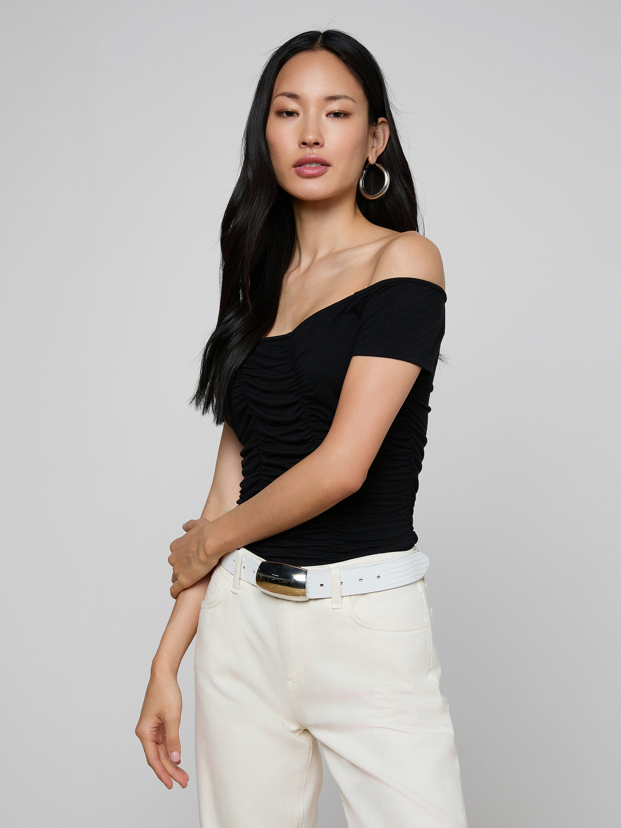 A woman with long dark hair stands against a plain light background, wearing the Baliegh Ruched Bodysuit—a black off-the-shoulder rayon stretch jersey top—paired with large hoop earrings and white high-waisted belted pants.