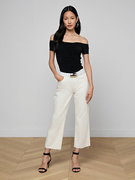 Woman wearing the Baliegh Ruched Bodysuit with an off-the-shoulder neckline, paired with white wide-leg pants, a black belt, and black high-heeled sandals, standing against a plain light background with her hands in her pockets.