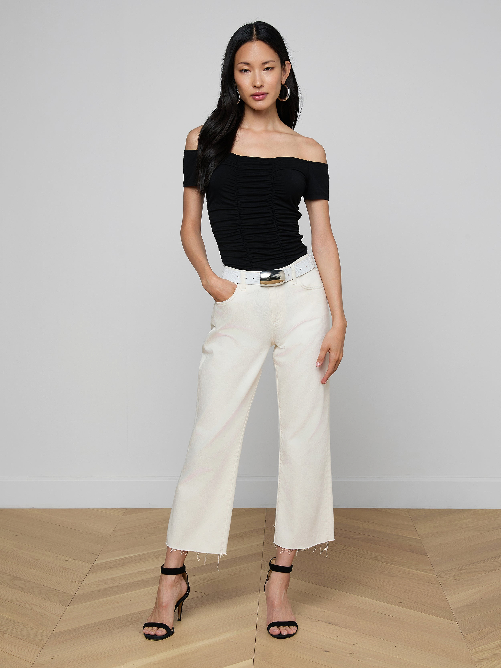 Woman wearing the Baliegh Ruched Bodysuit with an off-the-shoulder neckline, paired with white wide-leg pants, a black belt, and black high-heeled sandals, standing against a plain light background with her hands in her pockets.