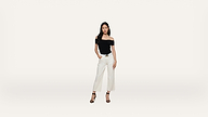 A woman stands against a plain white background, wearing a black off-the-shoulder top, white wide-leg pants, and black high-heeled sandals.