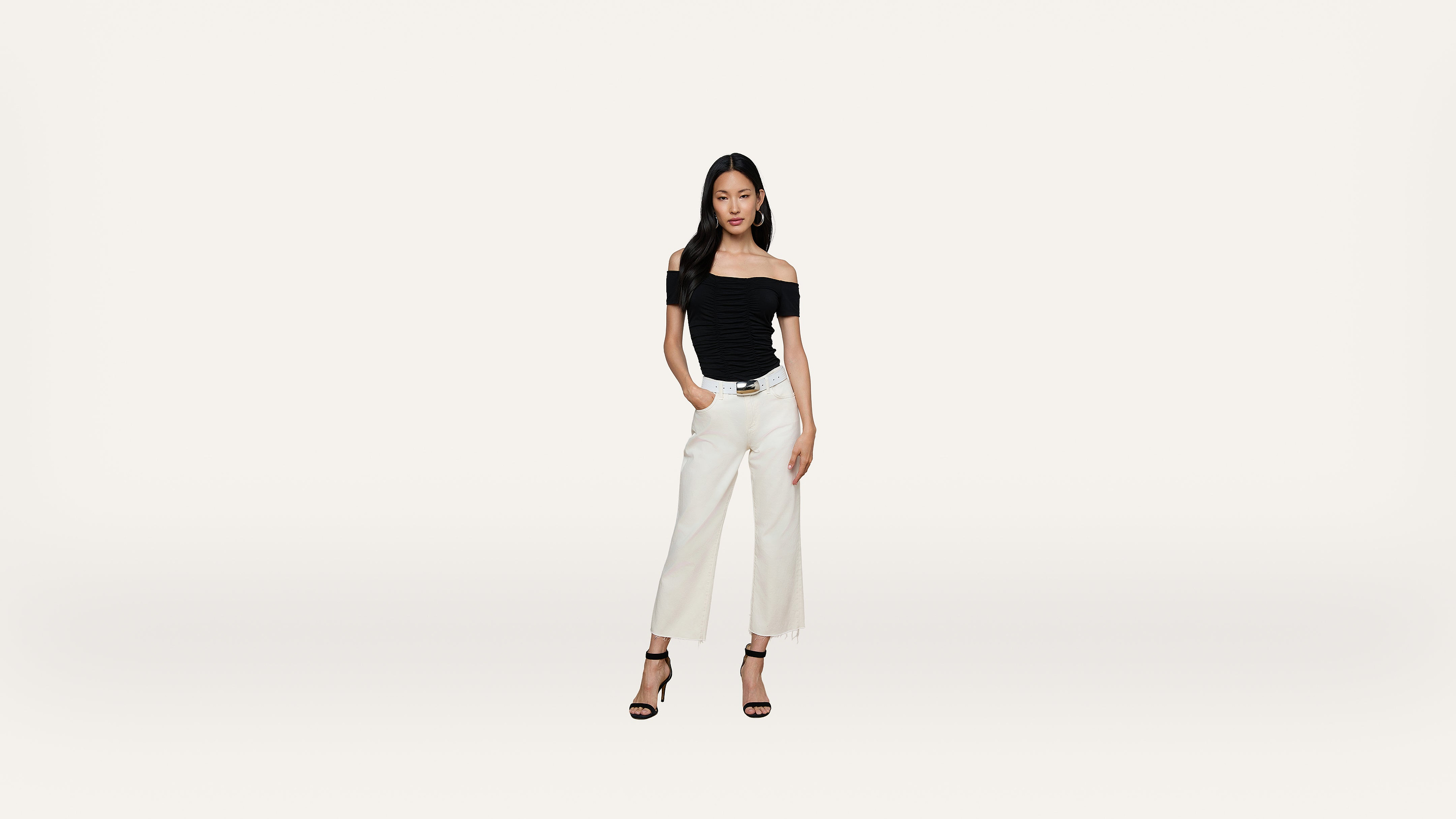 A woman stands against a plain white background, wearing a black off-the-shoulder top, white wide-leg pants, and black high-heeled sandals.