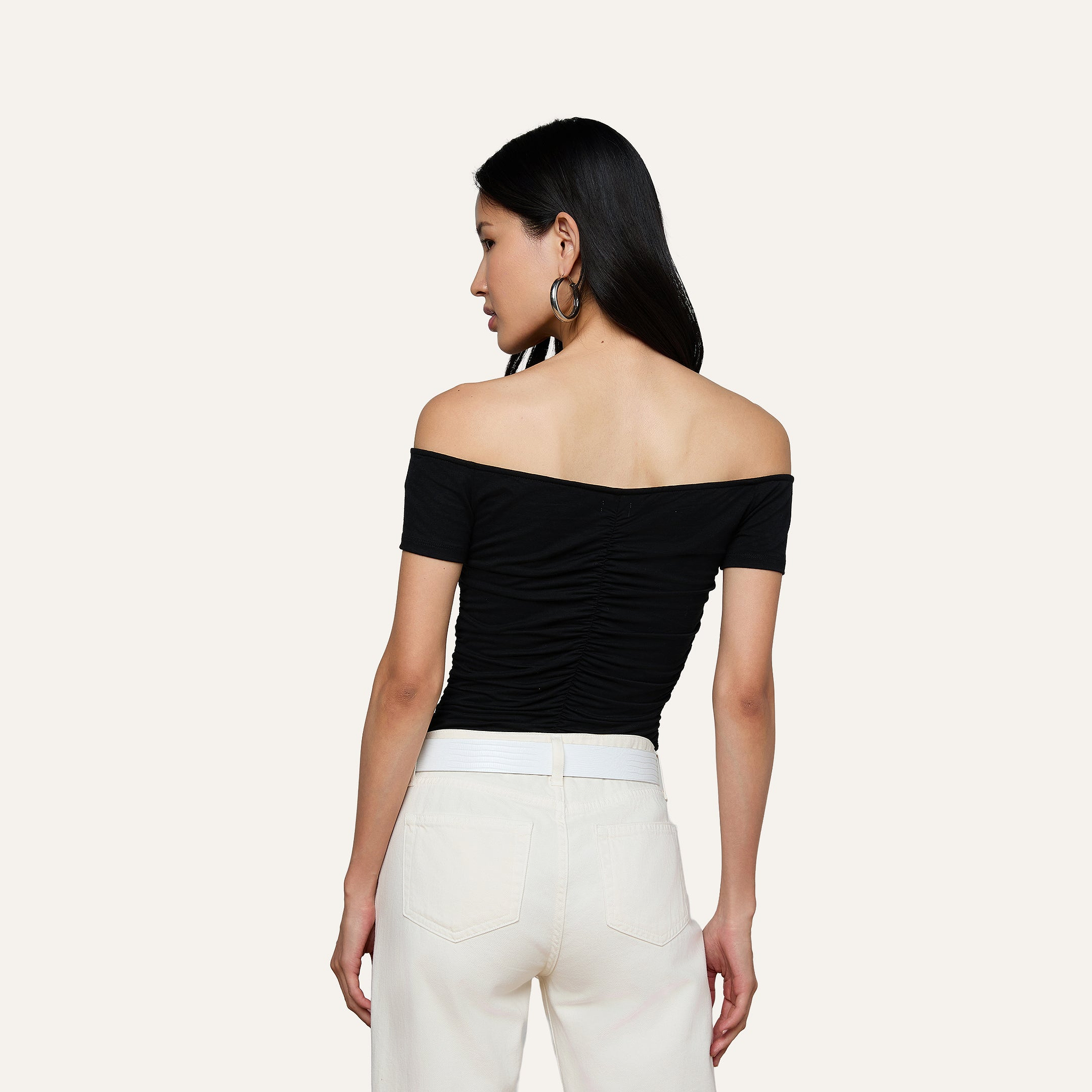 A woman stands with her back to the camera, wearing the Baliegh Ruched Bodysuit—a black off-the-shoulder piece crafted from rayon stretch jersey—paired with white pants. She has long dark hair, hoop earrings, and is set against a plain, light-colored background.