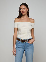 A woman wearing the Baliegh Ruched Bodysuit in vintage white, paired with blue jeans and a brown belt, stands with one hand in her pocket against a plain background.