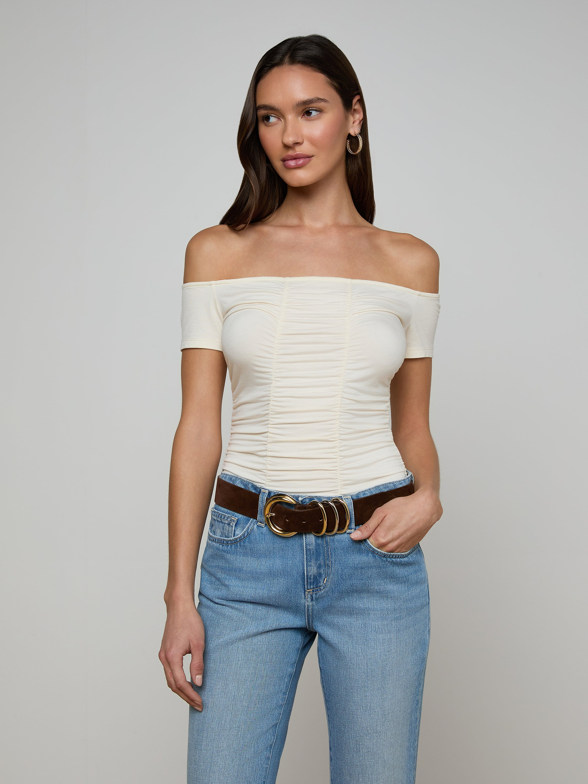 A woman wearing the Baliegh Ruched Bodysuit in vintage white, paired with blue jeans and a brown belt, stands with one hand in her pocket against a plain background.