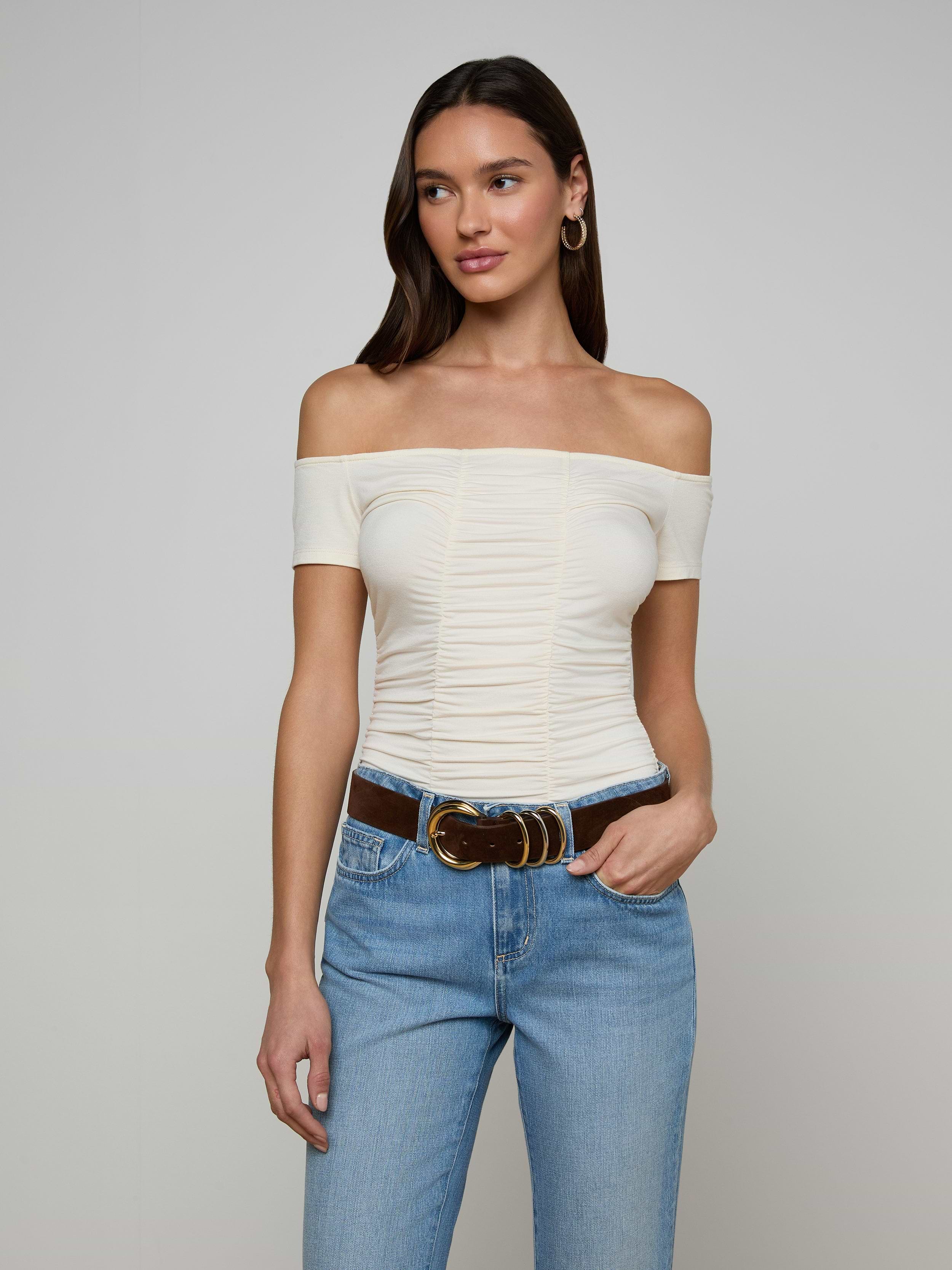 A woman wearing the Baliegh Ruched Bodysuit in vintage white, paired with blue jeans and a brown belt, stands with one hand in her pocket against a plain background.