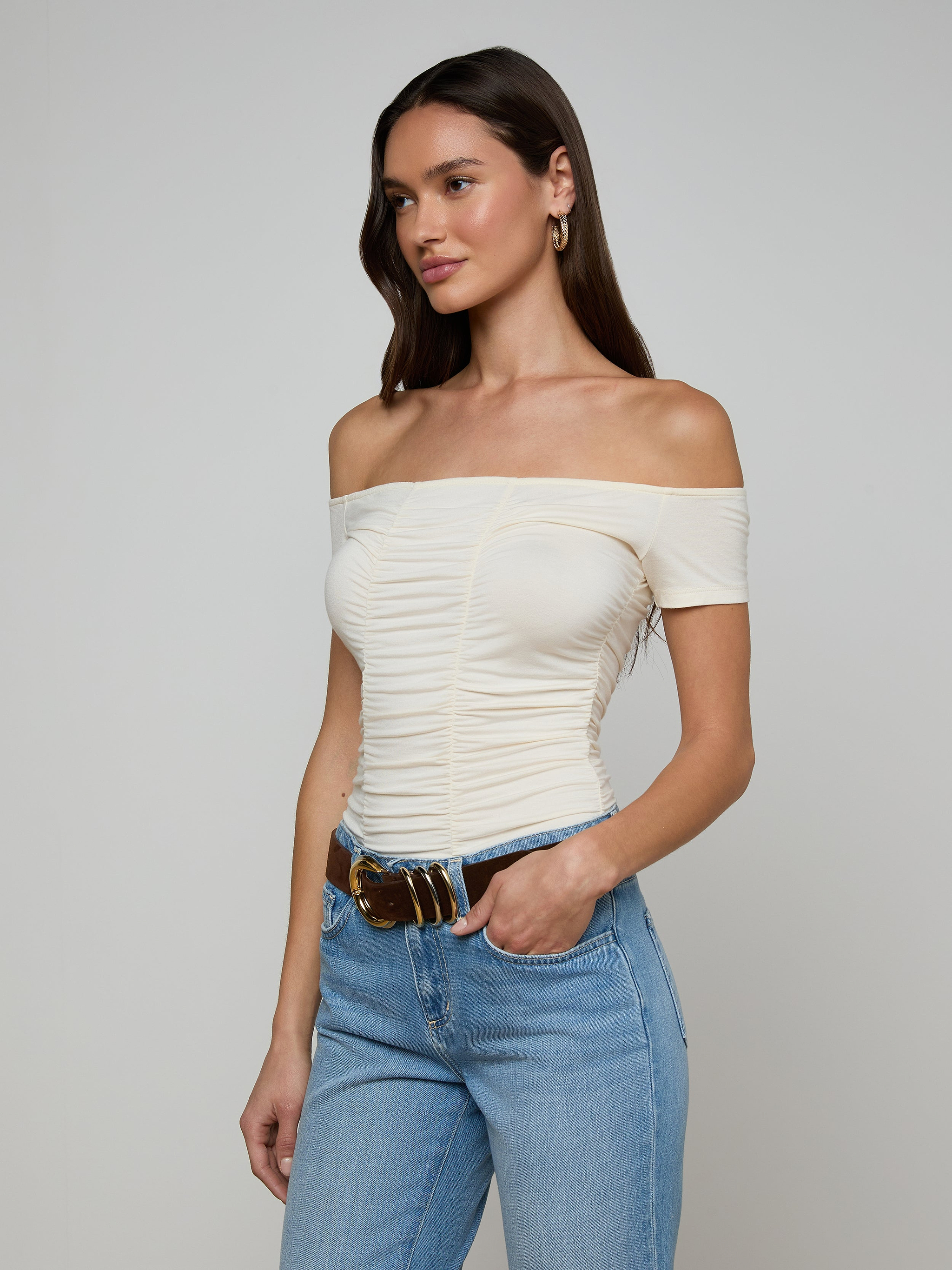 Woman wearing the Baliegh Ruched Bodysuit in vintage white, paired with light blue jeans, a brown belt, and gold hoop earrings, standing against a plain light background.