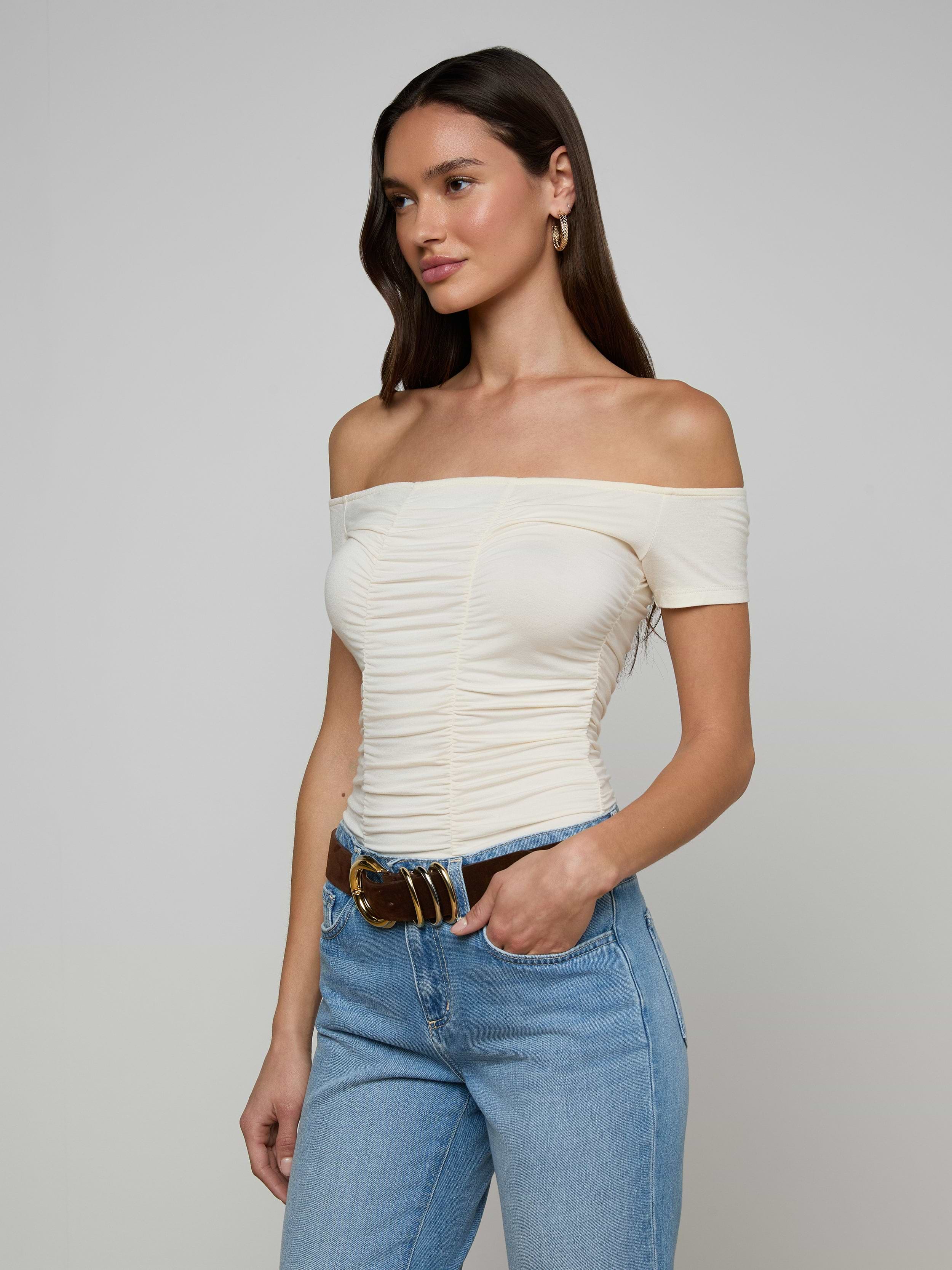 Woman wearing the Baliegh Ruched Bodysuit in vintage white, paired with light blue jeans, a brown belt, and gold hoop earrings, standing against a plain light background.