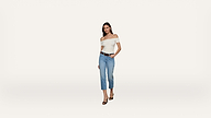 A woman stands against a plain white background, wearing a white off-the-shoulder top, blue cropped jeans, a black belt, and black sandals. She has one hand in her pocket and is facing forward, smiling slightly.