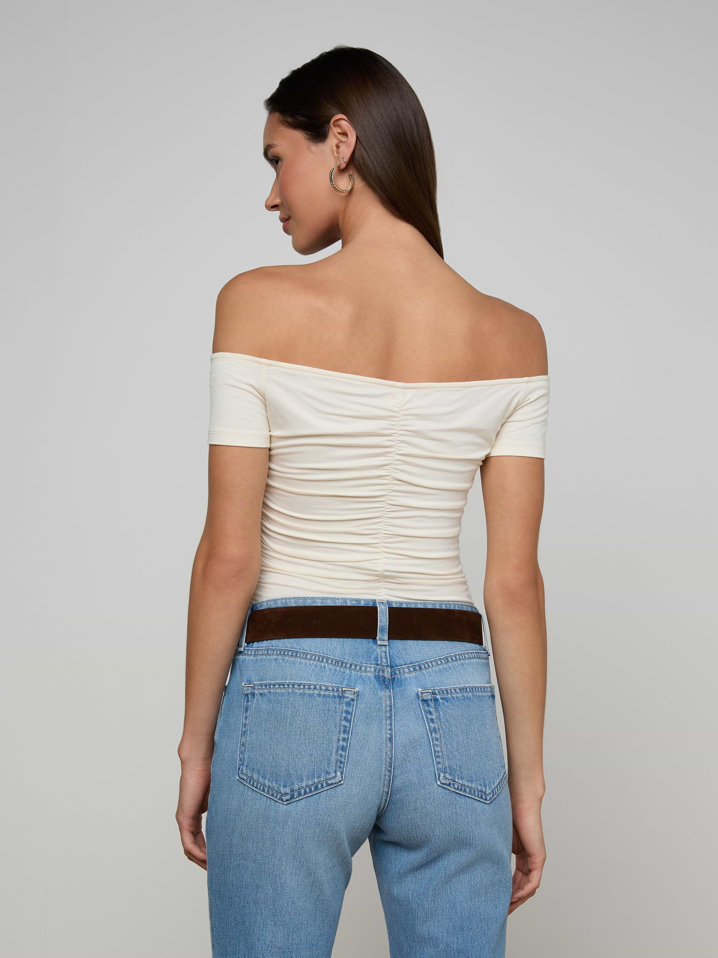 A woman is standing with her back to the camera, wearing the vintage-inspired white Baliegh Ruched Bodysuit and light blue jeans.