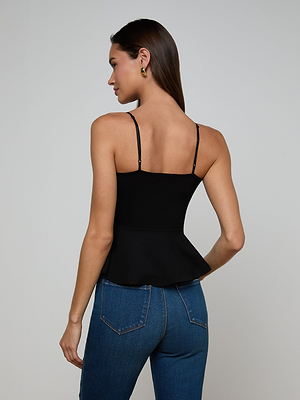 A woman with long brown hair stands with her back to the camera in a black Parisa Peplum Top and blue jeans. She wears gold hoop earrings, her left arm relaxed by her side, against a plain light gray background.