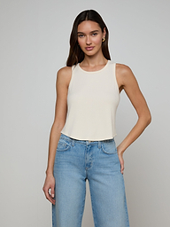 A woman stands against a plain off-white background, wearing the Cael Tank Top—an ultra-soft, sleeveless white crop top—paired with light blue jeans and her right hand in her pocket.