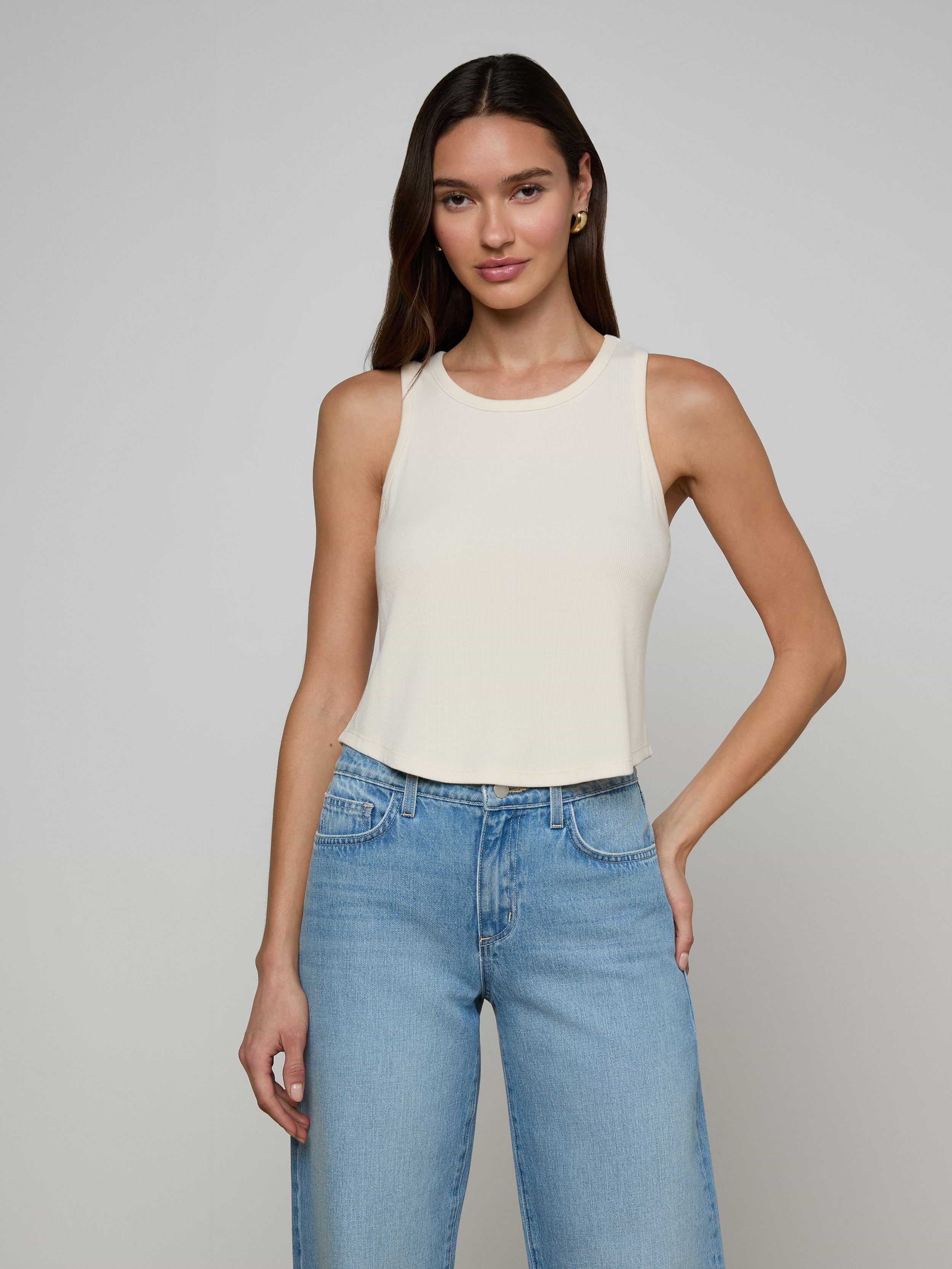 A woman stands against a plain off-white background, wearing the Cael Tank Top—an ultra-soft, sleeveless white crop top—paired with light blue jeans and her right hand in her pocket.