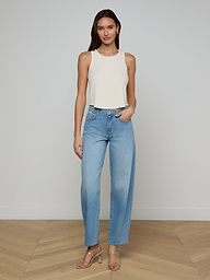 A woman stands against a plain white background, wearing the Cael Tank Top in white, paired with light blue high-waisted jeans and tan heeled sandals. She has long hair and poses relaxedly with one hand in her pocket.