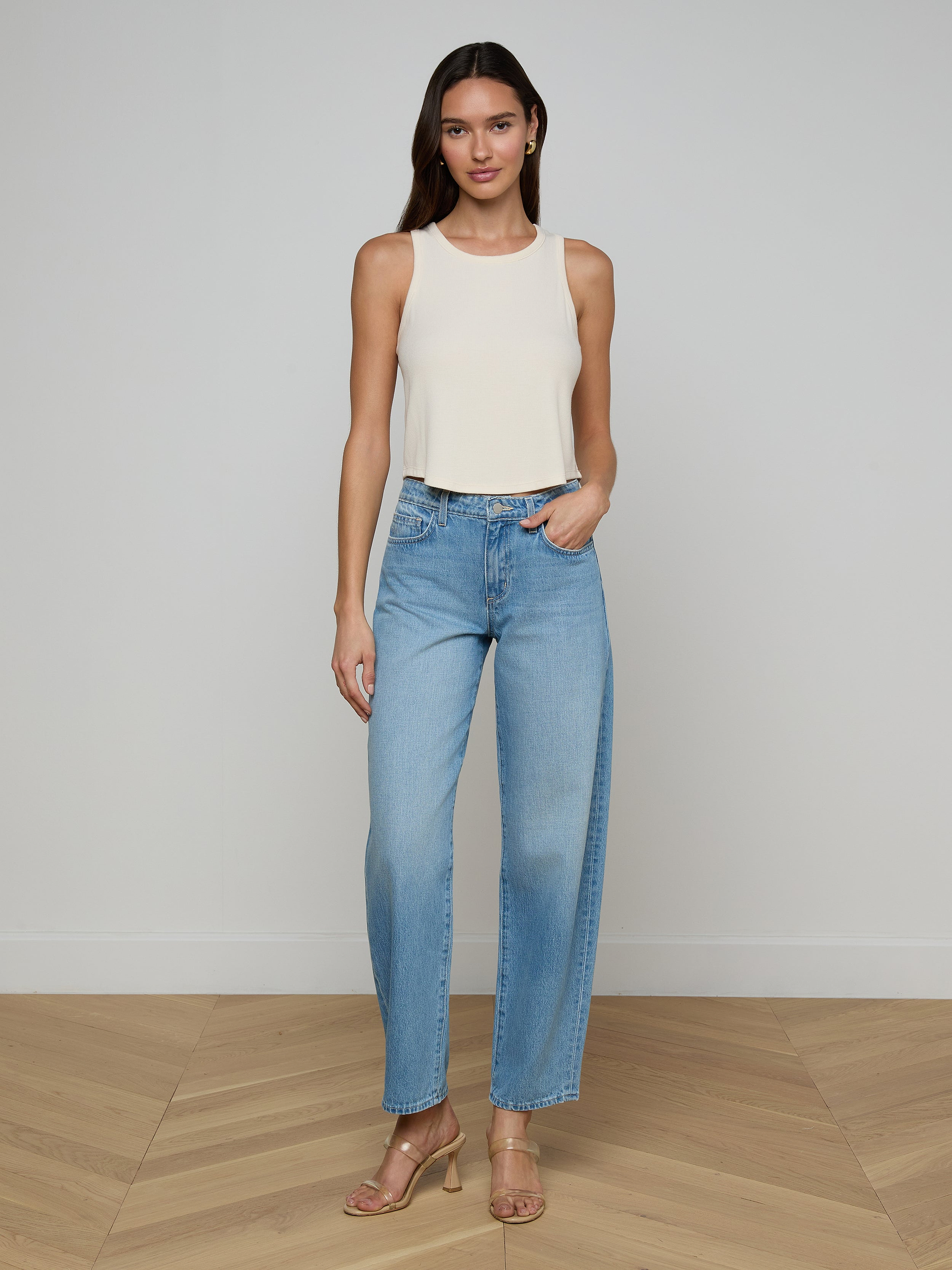 A woman stands against a plain white background, wearing the Cael Tank Top in white, paired with light blue high-waisted jeans and tan heeled sandals. She has long hair and poses relaxedly with one hand in her pocket.