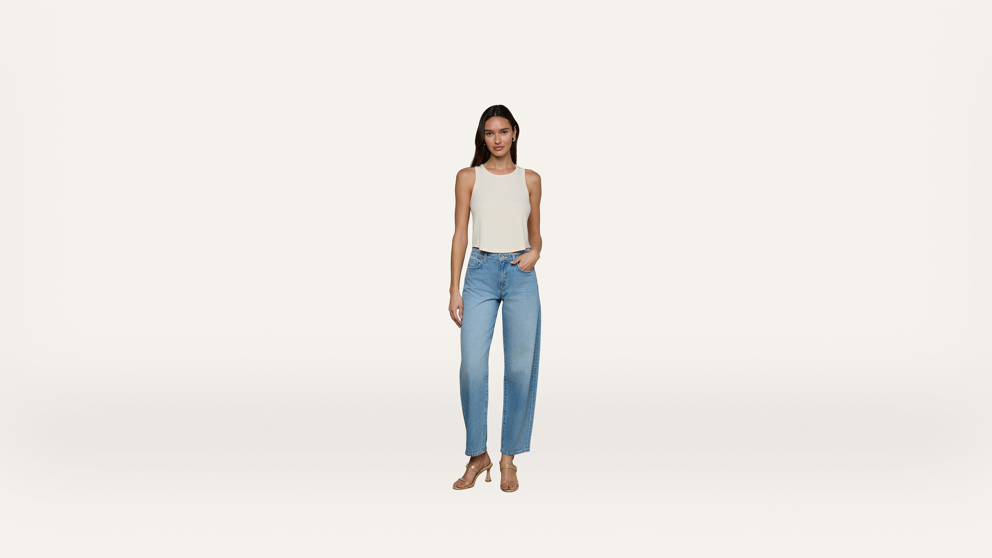 A woman stands facing forward, wearing a white sleeveless top, light blue jeans, and beige heels, with one hand in her pocket and a neutral expression on a plain white background.