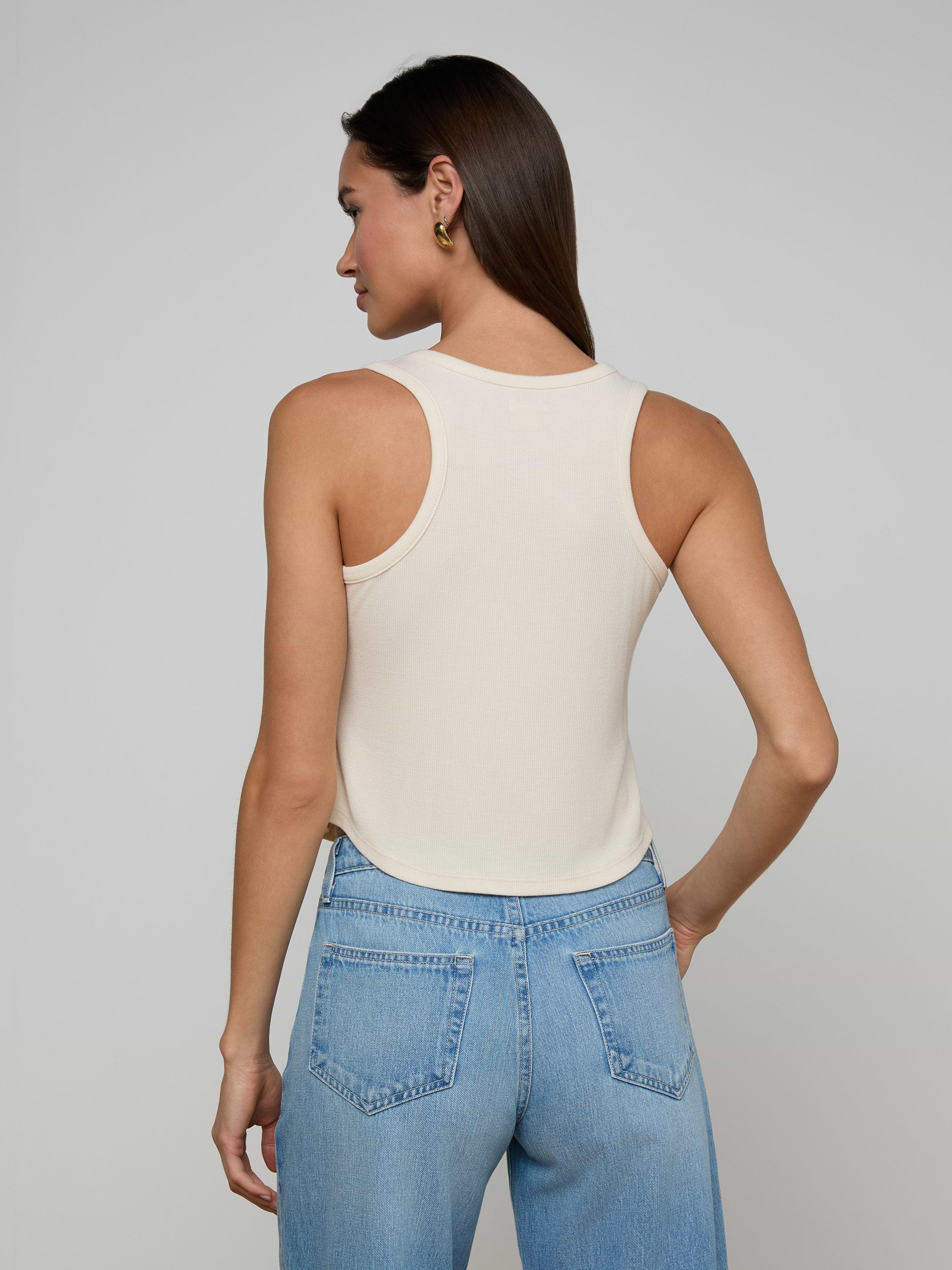 A woman stands with her back to the camera, wearing the Cael Tank Top in white and light blue jeans, set against a plain light background.