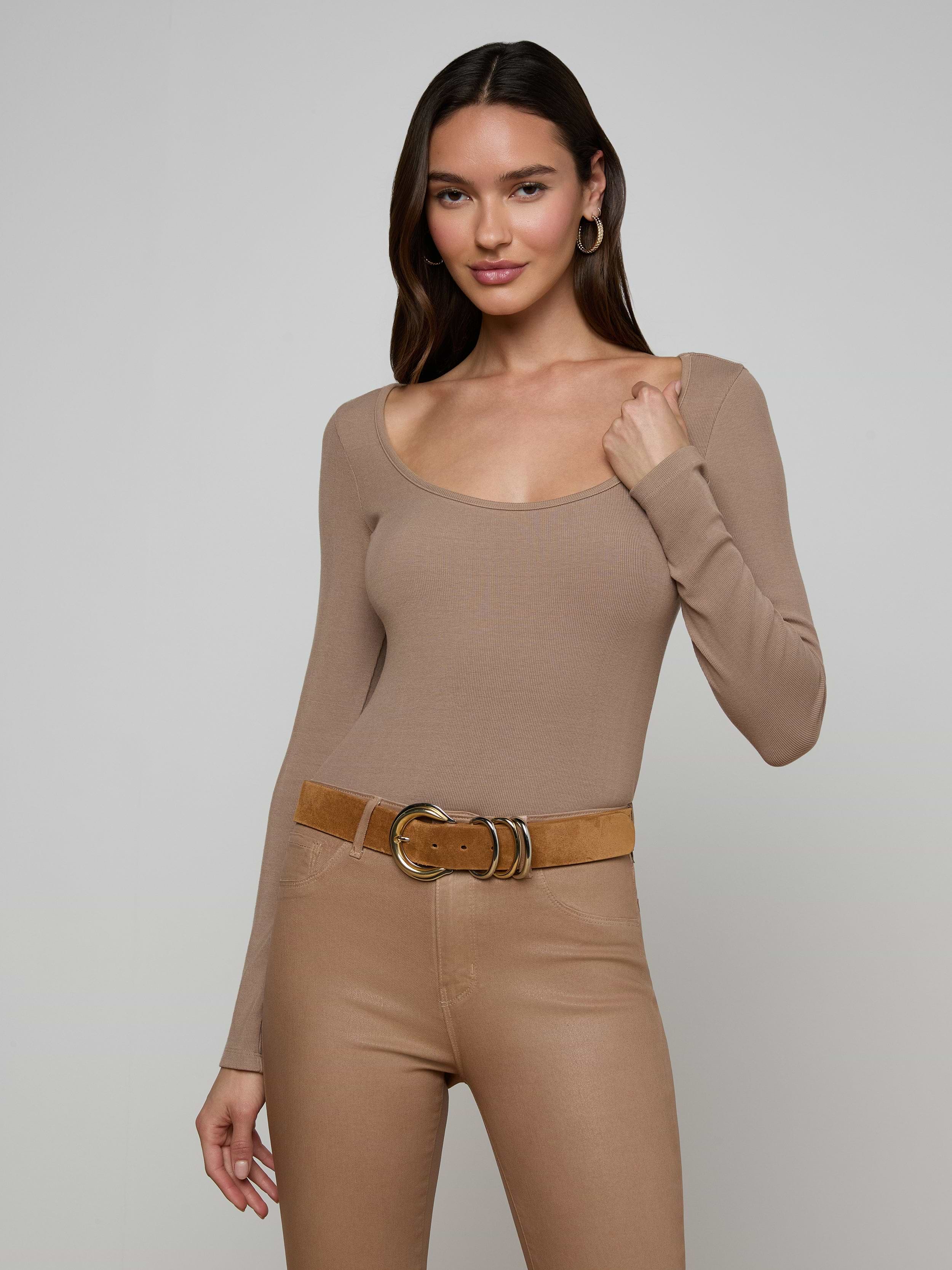 Wearing the Vanya Scoopneck Top in taupe, this woman has it tucked into high-waisted beige pants paired with a wide brown belt, standing against a plain light background and smiling softly.