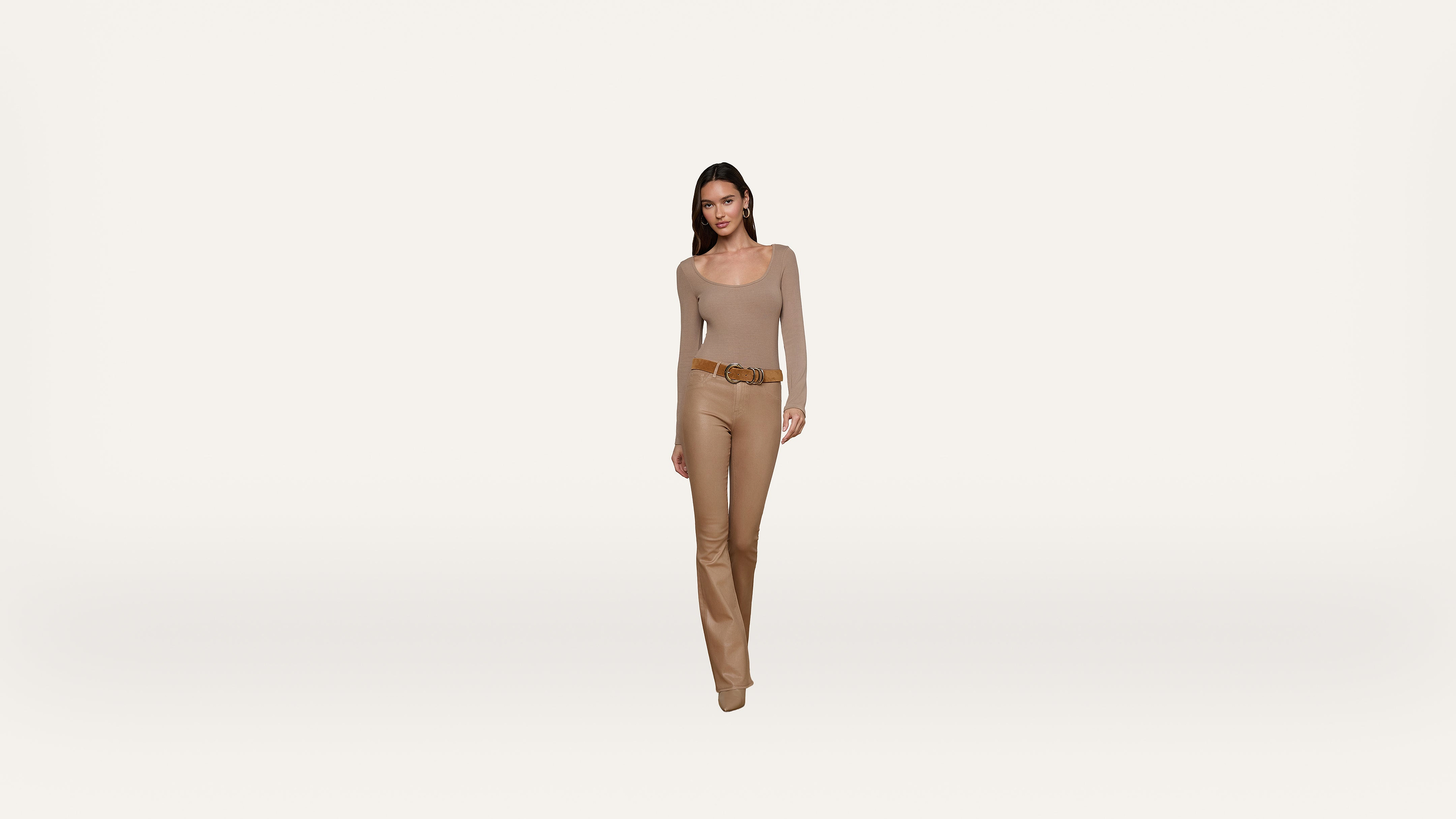 A woman stands against a plain white background wearing a beige long-sleeve top, matching pants, and a brown belt. She has straight dark hair and is looking forward with a neutral expression.