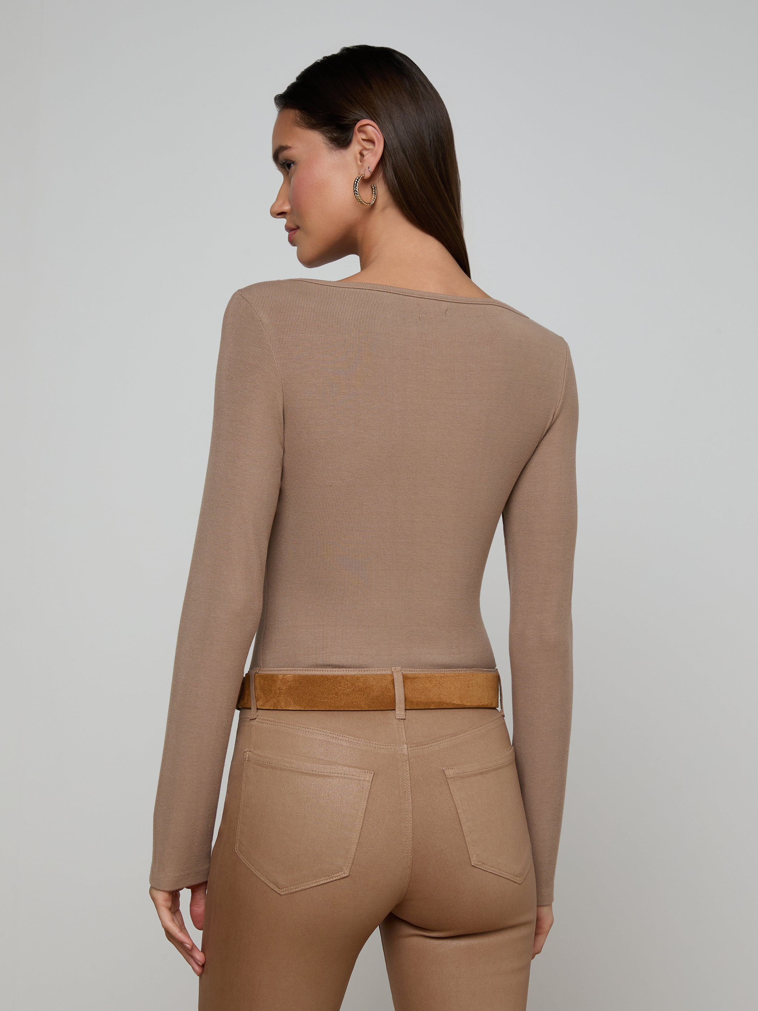 The woman, seen from behind, is wearing the Vanya Scoopneck Top—a fitted, long-sleeve style made from soft stretch rib—tucked into high-waisted tan pants with back pockets and a brown belt. Her straight hair falls loose as she stands against a plain light background.