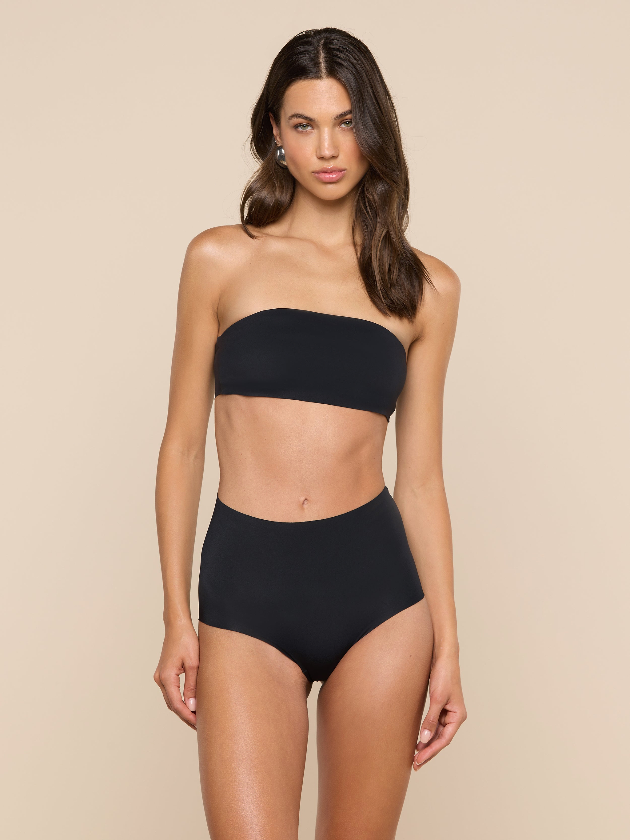 A woman stands against a plain light background, wearing a black Mietta Knit Bandeau Top and high-waisted bikini bottoms. She has long brown hair and a neutral expression.