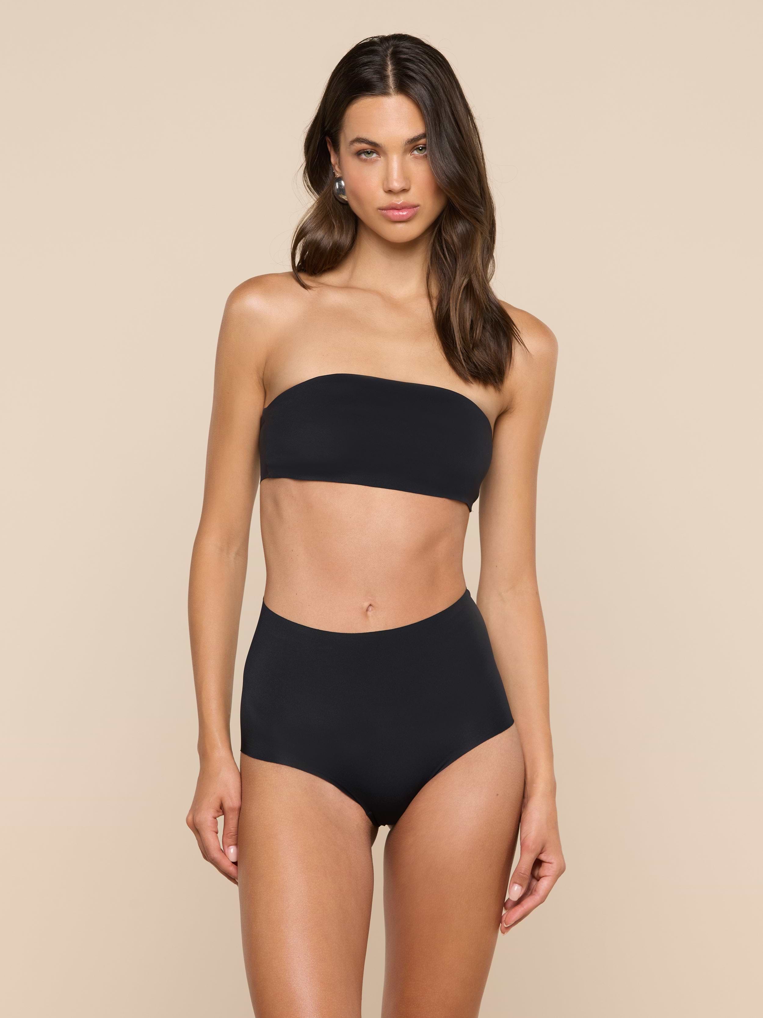 A woman stands against a plain light background, wearing a black Mietta Knit Bandeau Top and high-waisted bikini bottoms. She has long brown hair and a neutral expression.