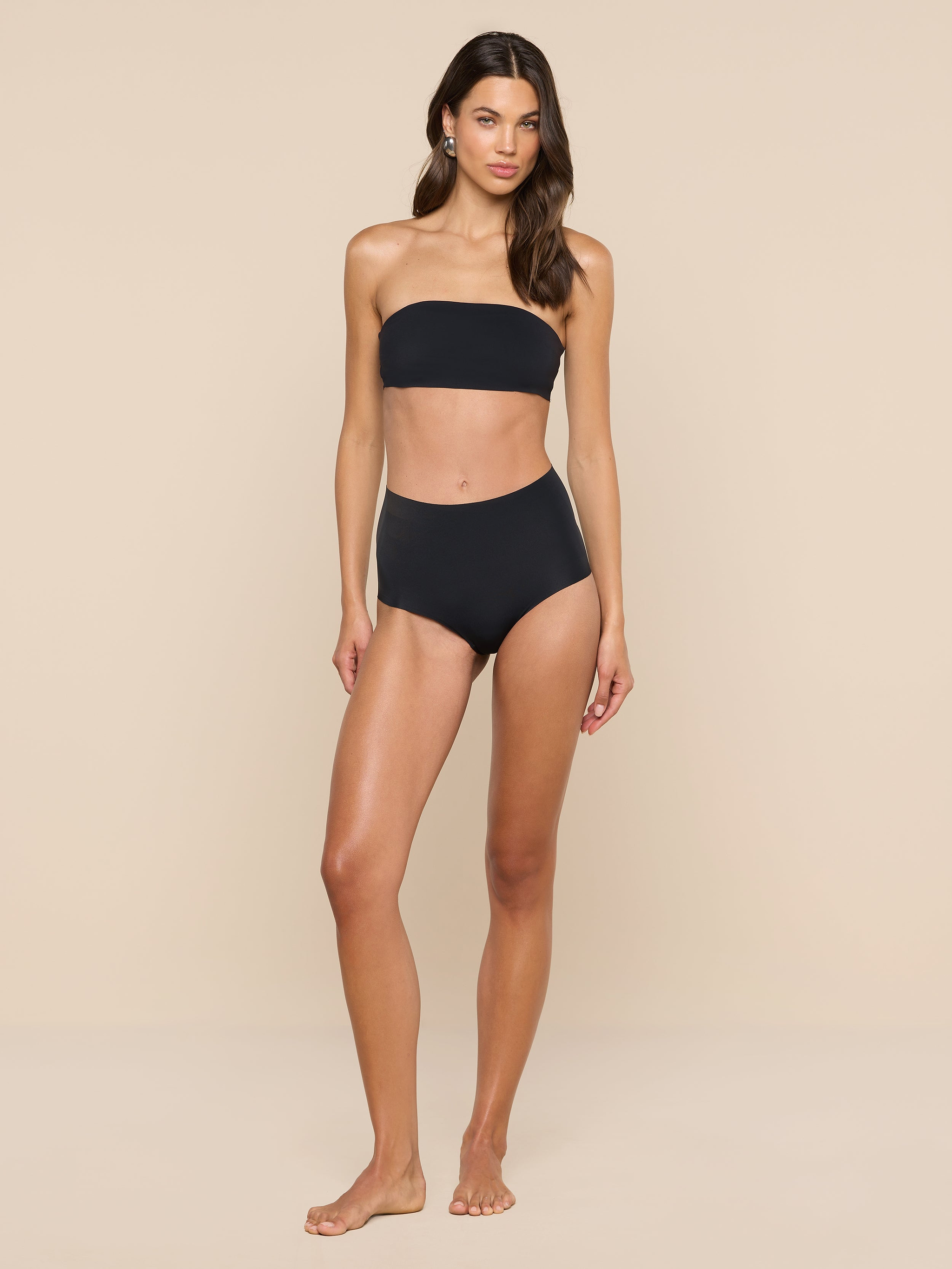 A woman stands against a plain white background, wearing the supportive Mietta Knit Bandeau Top in black paired with high-waisted bikini bottoms.