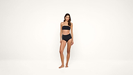 A woman stands barefoot against a plain white background, wearing a black bandeau bikini.