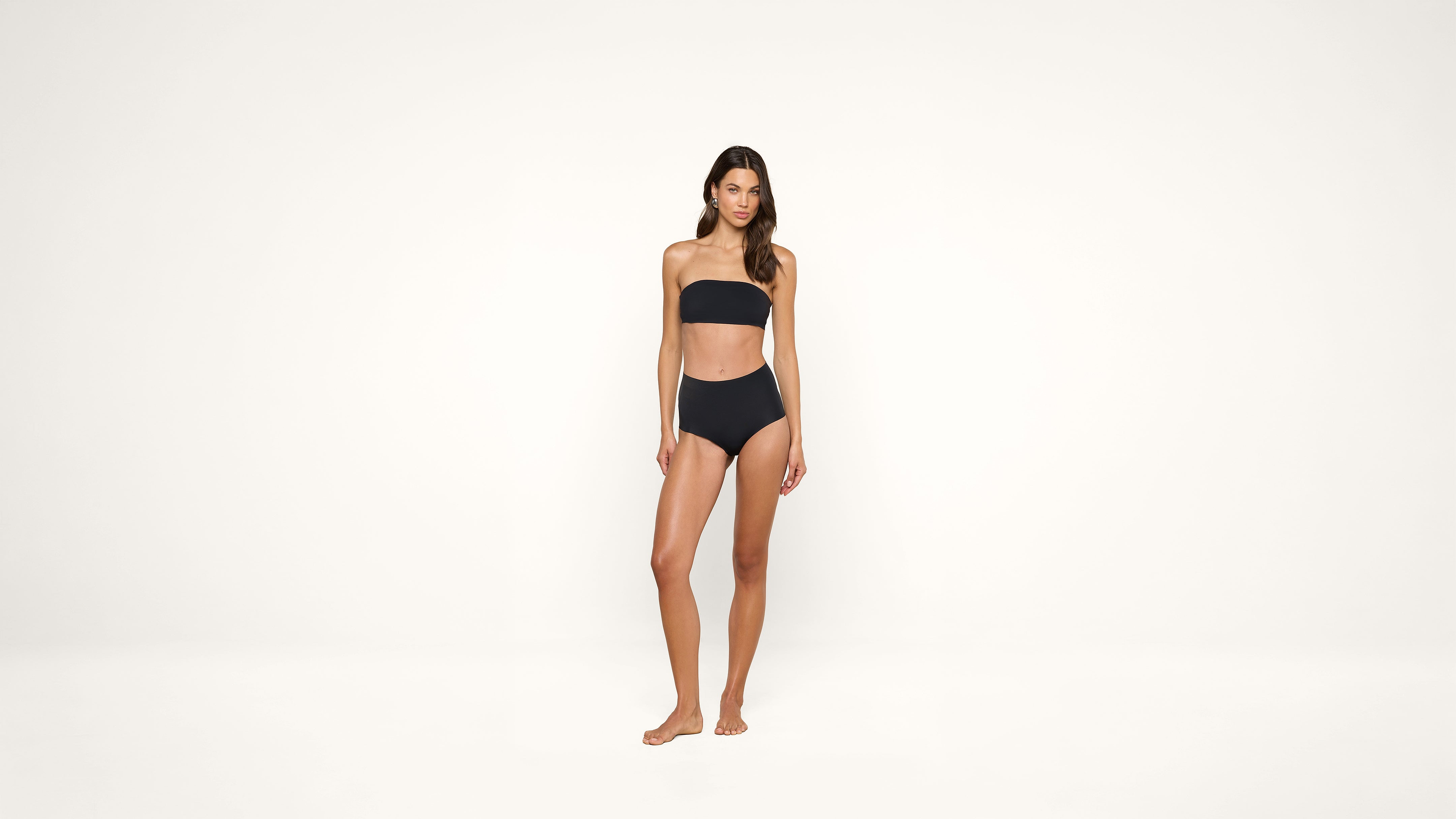 A woman stands barefoot against a plain white background, wearing a black bandeau bikini.