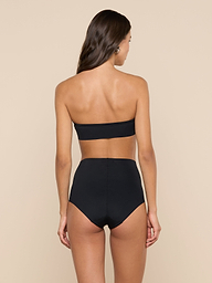 A woman with long brown hair stands with her back to the camera against a plain, light background, wearing the black Mietta Knit Bandeau Top paired with high-waisted bottoms.