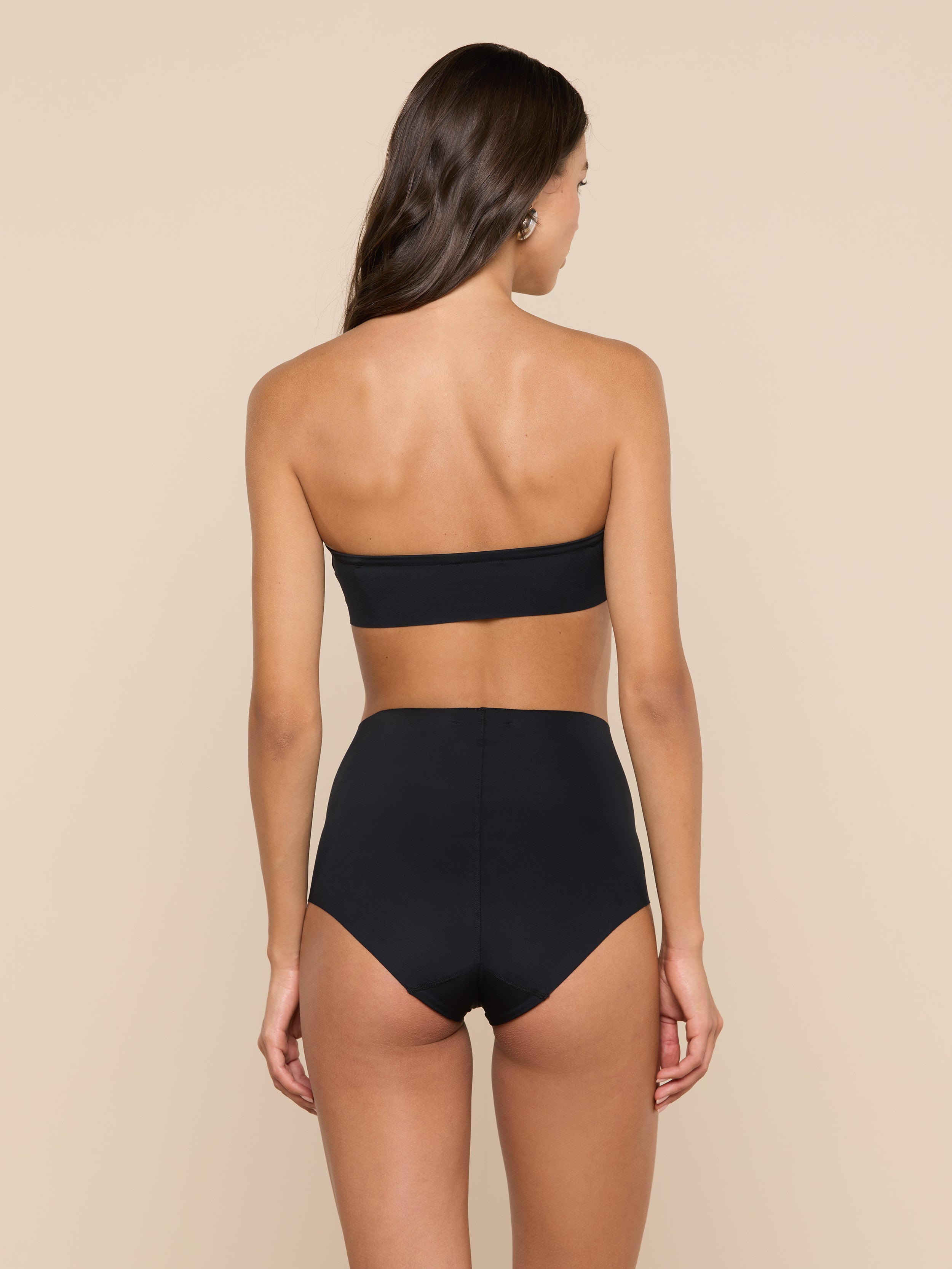 A woman with long brown hair stands with her back to the camera against a plain, light background, wearing the black Mietta Knit Bandeau Top paired with high-waisted bottoms.