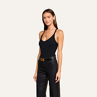 A woman with long brown hair wears the Wrenna Racerback Bodysuit— a black fitted ribbed tank bodysuit—paired with black leather pants, gold jewelry, and a black belt, standing against a plain beige background.