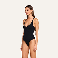A woman with long brown hair wears the Wrenna Racerback Bodysuit, a black fitted tank style, against a plain beige background. She is facing slightly to the left, looking forward with a neutral expression.