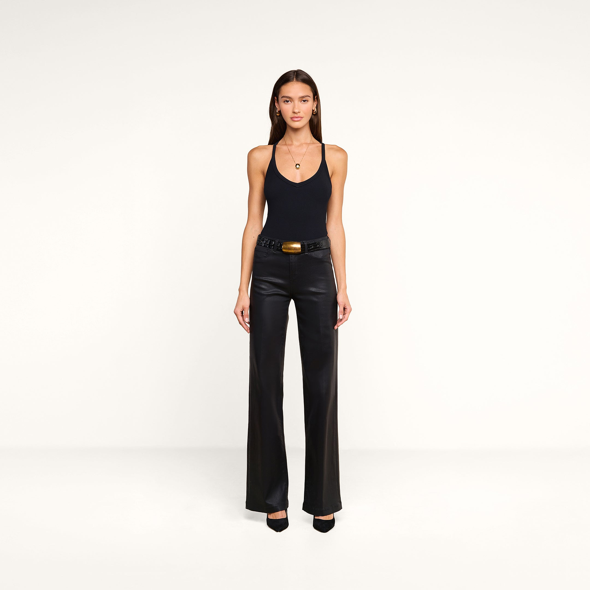 A woman with long brown hair and a neutral expression stands against a beige background, wearing the Wrenna Racerback Bodysuit in fitted rib fabric. She pairs it with black wide-leg leather pants featuring a chunky belt, black heels, gold hoop earrings, and a necklace.
