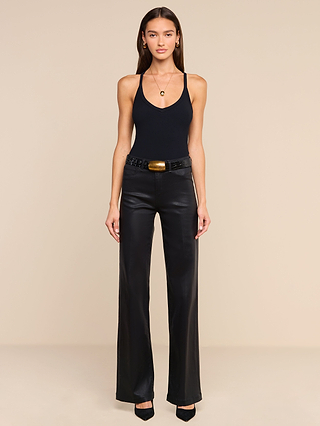 A woman with long brown hair and a neutral expression stands against a beige background, wearing the Wrenna Racerback Bodysuit in fitted rib fabric. She pairs it with black wide-leg leather pants featuring a chunky belt, black heels, gold hoop earrings, and a necklace.
