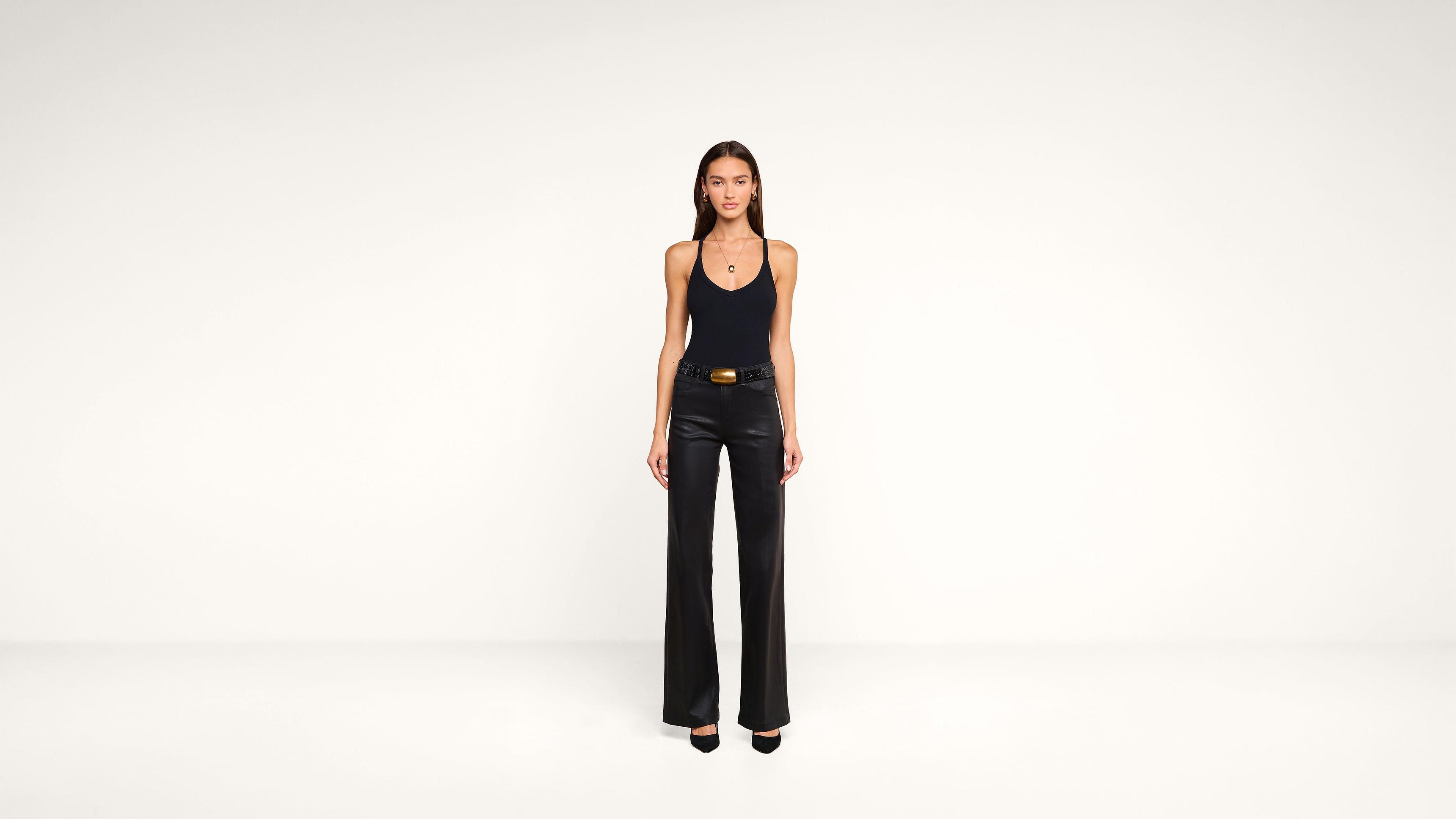 A woman stands facing forward in a minimal studio, wearing a black tank top, black wide-leg pants, a belt, and black heels. The background is plain white.