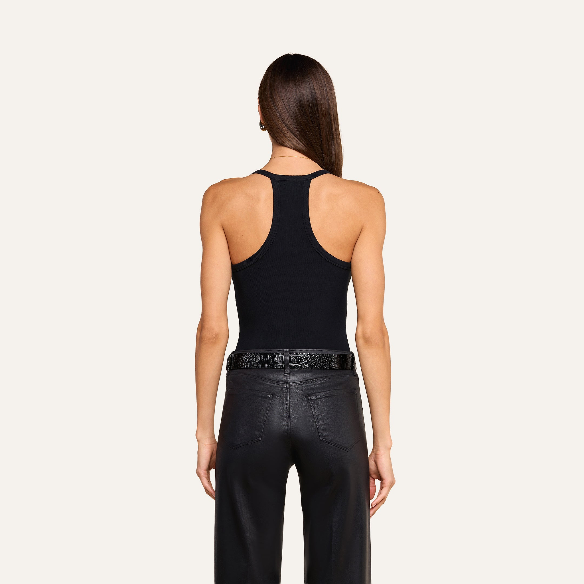 A woman with long brown hair is shown from behind, wearing the Wrenna Racerback Bodysuit in black rib fabric and black leather pants with a textured belt, standing against a beige background.