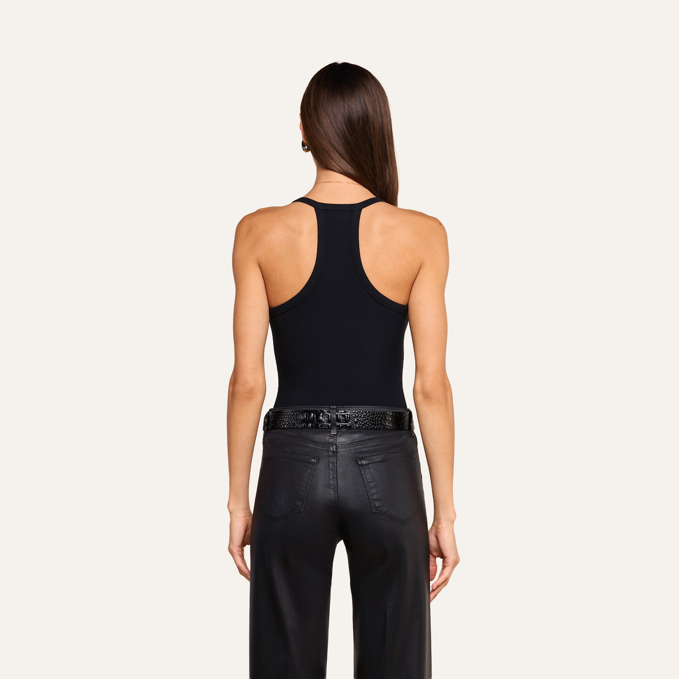 A woman with long brown hair is shown from behind, wearing the Wrenna Racerback Bodysuit in black rib fabric and black leather pants with a textured belt, standing against a beige background.