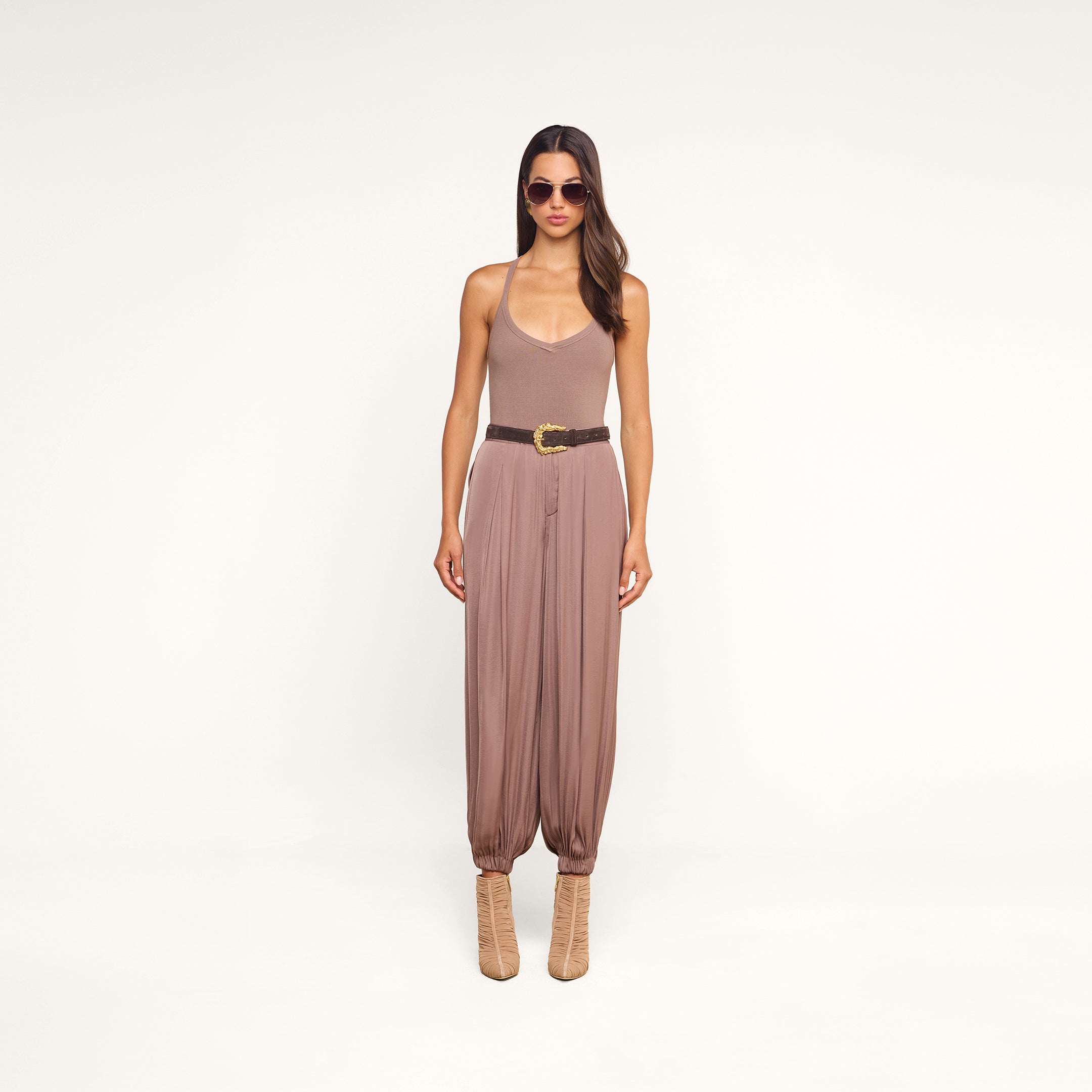A woman wearing sunglasses, the Wrenna Racerback Bodysuit in taupe rib fabric, loose mauve pants with a belt, and beige boots stands against a plain white background.