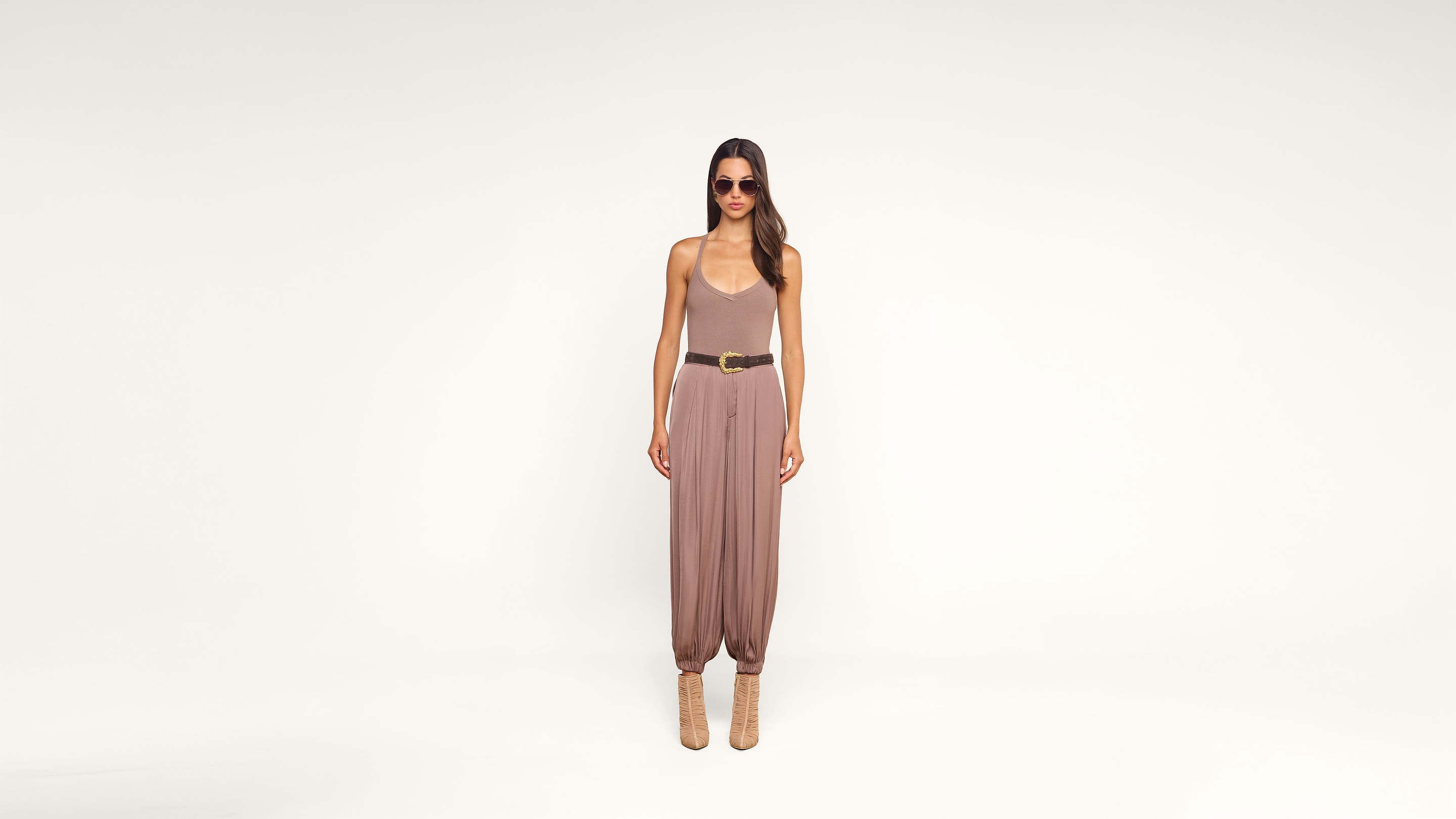 Woman in sunglasses wearing a tan jumpsuit with a brown belt and ankle boots stands against a plain white background.