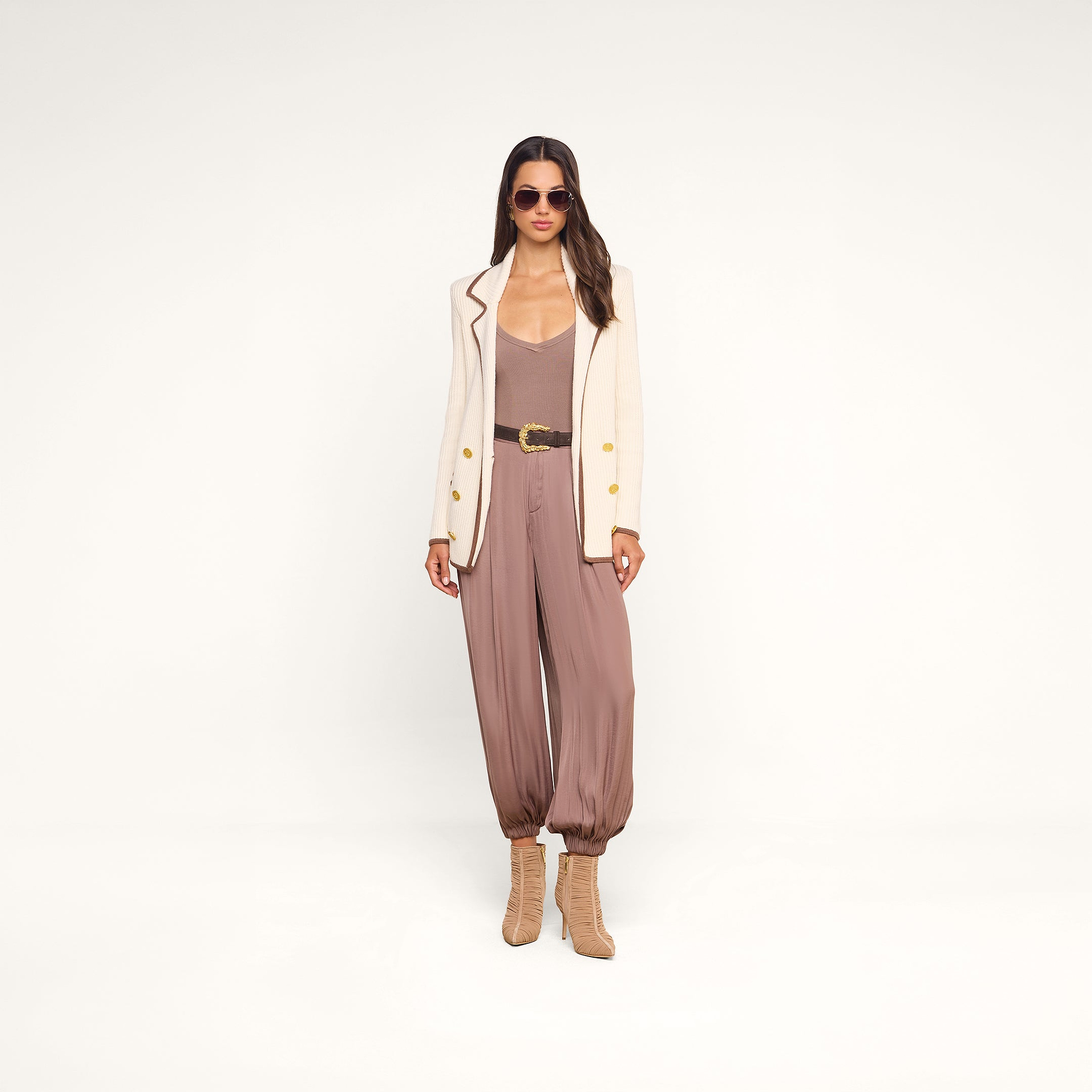Woman wearing sunglasses, a cream blazer layered over the Wrenna Racerback Bodysuit in taupe rib fabric, brown belt, loose mauve pants gathered at the ankles, and tan heeled boots stands against a plain white background.