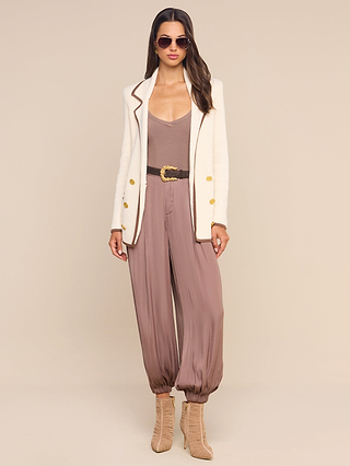 Woman wearing sunglasses, a white blazer with gold buttons, the Wrenna Racerback Bodysuit crafted from taupe rib fabric and cinched with a belt, paired with beige lace-up heels, standing against a plain beige background.