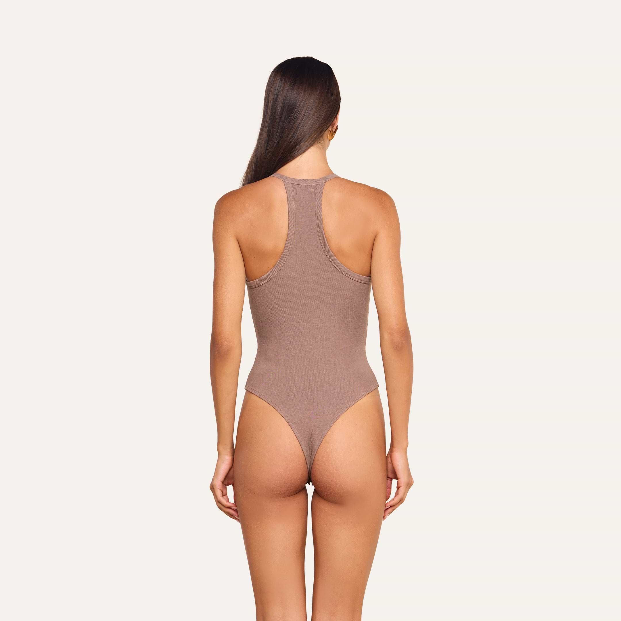 A woman stands facing away, wearing the Wrenna Racerback Bodysuit in taupe rib fabric against a plain off-white background.