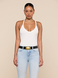 A woman stands facing forward against a beige background, wearing the Wrenna Racerback Bodysuit in white rib fabric, paired with light blue jeans and a black belt with a large gold buckle. Her hair is pulled back and she accessorizes with gold earrings.