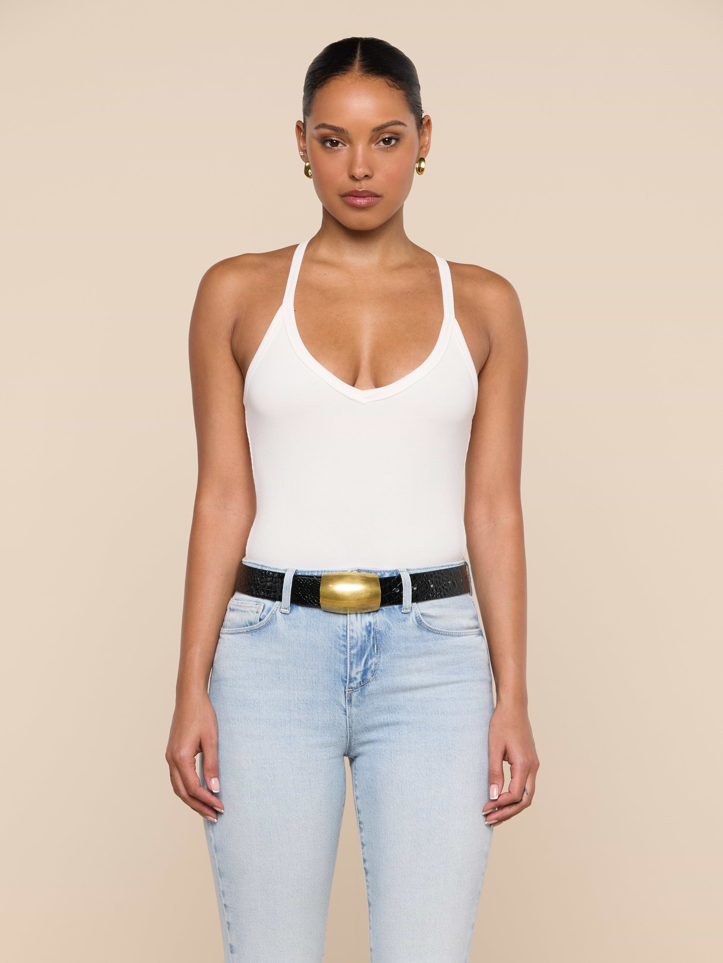 A woman stands facing forward against a beige background, wearing the Wrenna Racerback Bodysuit in white rib fabric, paired with light blue jeans and a black belt with a large gold buckle. Her hair is pulled back and she accessorizes with gold earrings.