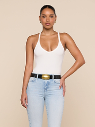 A woman stands against a beige background, wearing the Wrenna Racerback Bodysuit in white rib fabric, light blue jeans, a black belt with a gold buckle, and gold earrings. Her hair is pulled back and one hand rests on her hip.